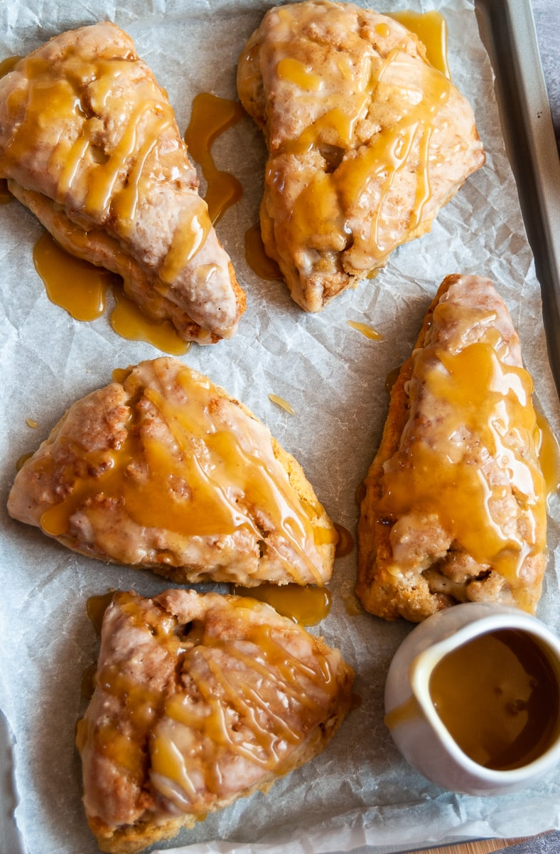 five apple and cinnamon scones drizzled with a caramel glaze on a baking tray and a small jug of caramel sauce