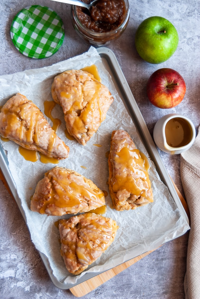 5 apple and cinnamon scones with a salted caramel glaze on a baking tray, a jar of apple butter with a spoon, a small jug of caramel sauce and a red and green apple
