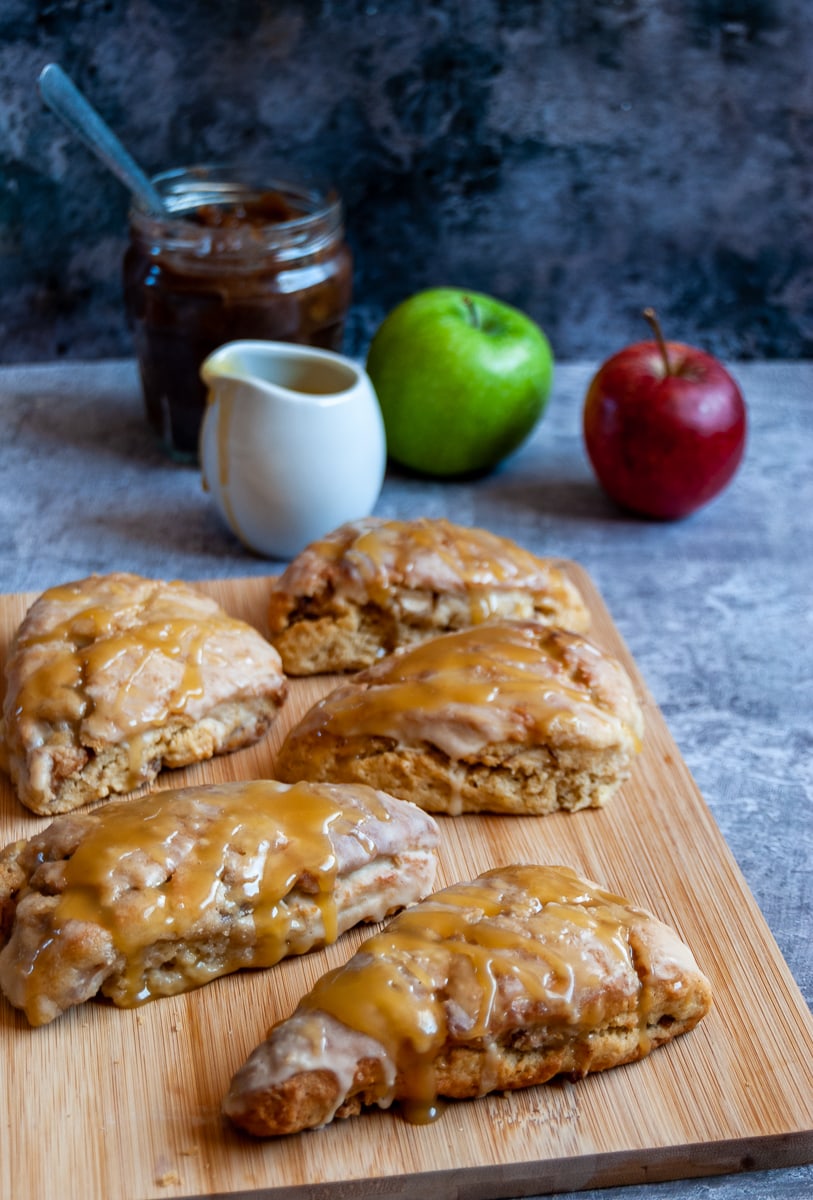 5 apple and cinnamon scones drizzled with a caramel glaze on a wooden board, a small jug of caramel sauce, a jar of apple butter and a red and green apple.
