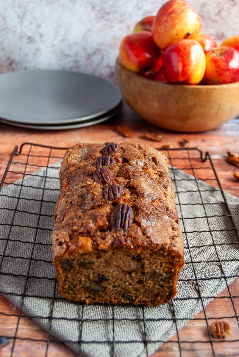 a courgette and apple loaf cake topped with pecan nuts on a black wire rack and grey napkin, two grey plates and a wooden bowl of red apples on a distressed looking wooden table