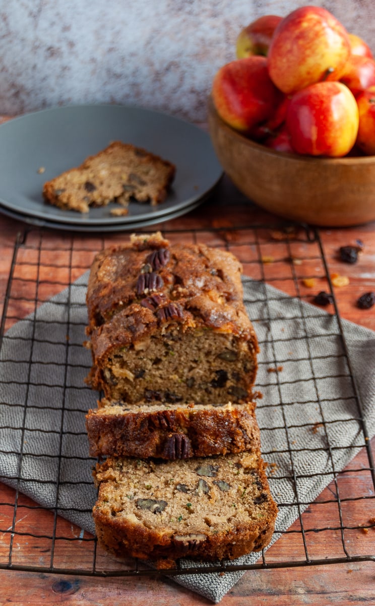 A courgette and apple loaf cake on a square black cooling rack, a grey napkin, a grey plate with a piece of the cake and a wooden bowl of red apples