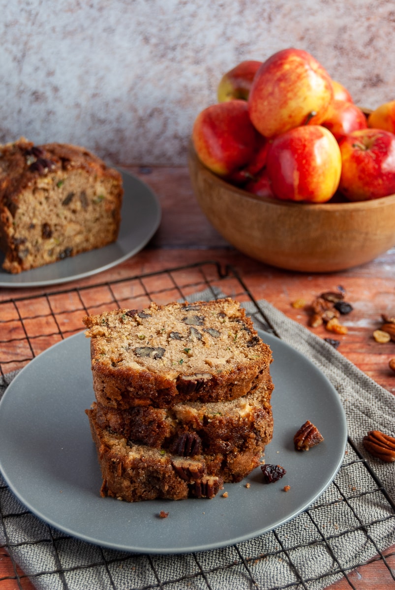 three slices of courgette and pecan bread on a grey plate/black cooling rack and a wooden bowl of red apples in the background.