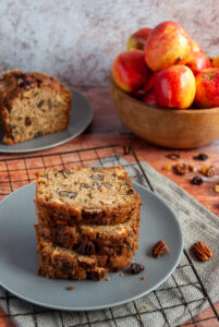 three slices of courgette and apple bread on a grey plate/black cooking rack and a wooden bowl of apples in the background