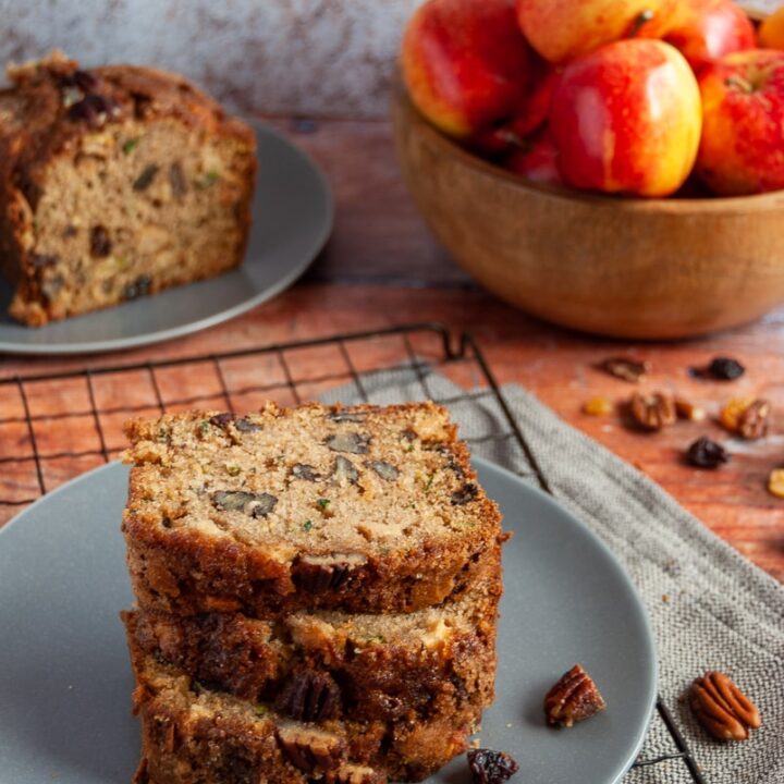 three slices of courgette and apple bread on a grey plate/black cooking rack and a wooden bowl of apples in the background