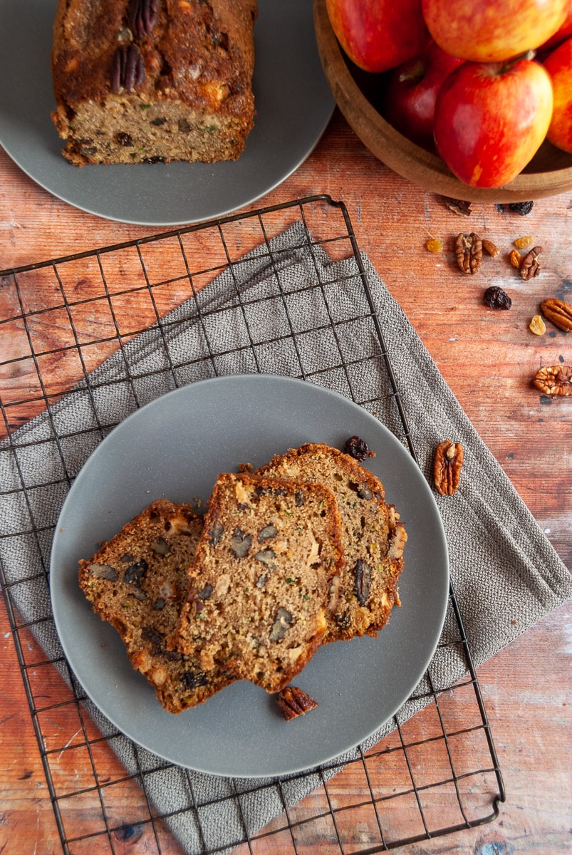 A flat lay photo of three slices of courgette and apple loaf cake on a grey plate and black wire rack. Pecan nuts and raisins are scattered around the plate along with a wooden bowl of red apples