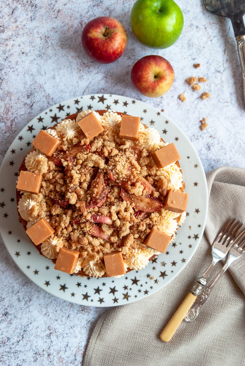 A flatly photo of an apple cake with custard buttercream, Crumble pieces and Fudge chunks on a white and gold star plate, two forks and a linen napkin and fresh apples