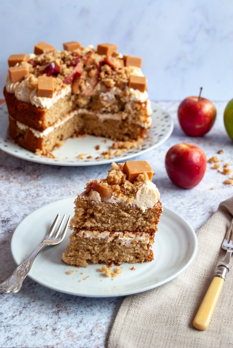 An slice of apple Crumble cake with custard buttercream and topped with Fudge on a white Plate with a cake fork, a large apple cake and red and green apples in the background