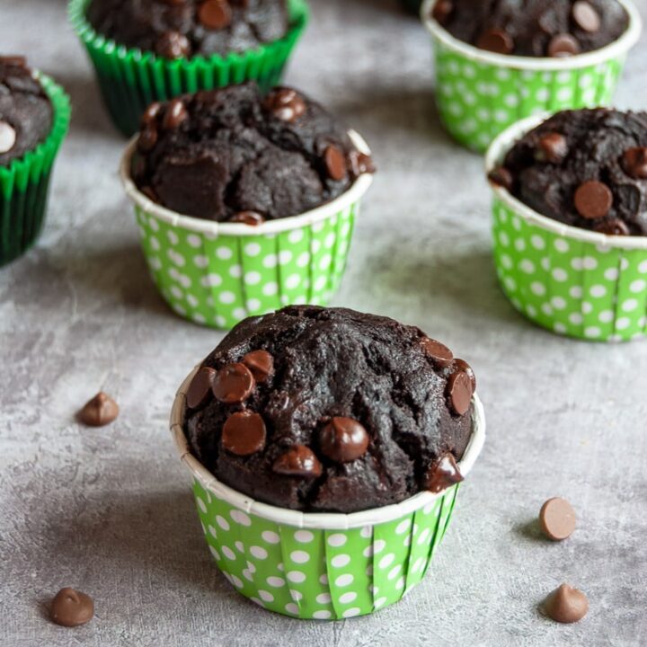 a batch of chocolate zucchini muffins with chocolate chips in green and white spotted muffin cases. chocolate chips are sprinkled all around the muffins