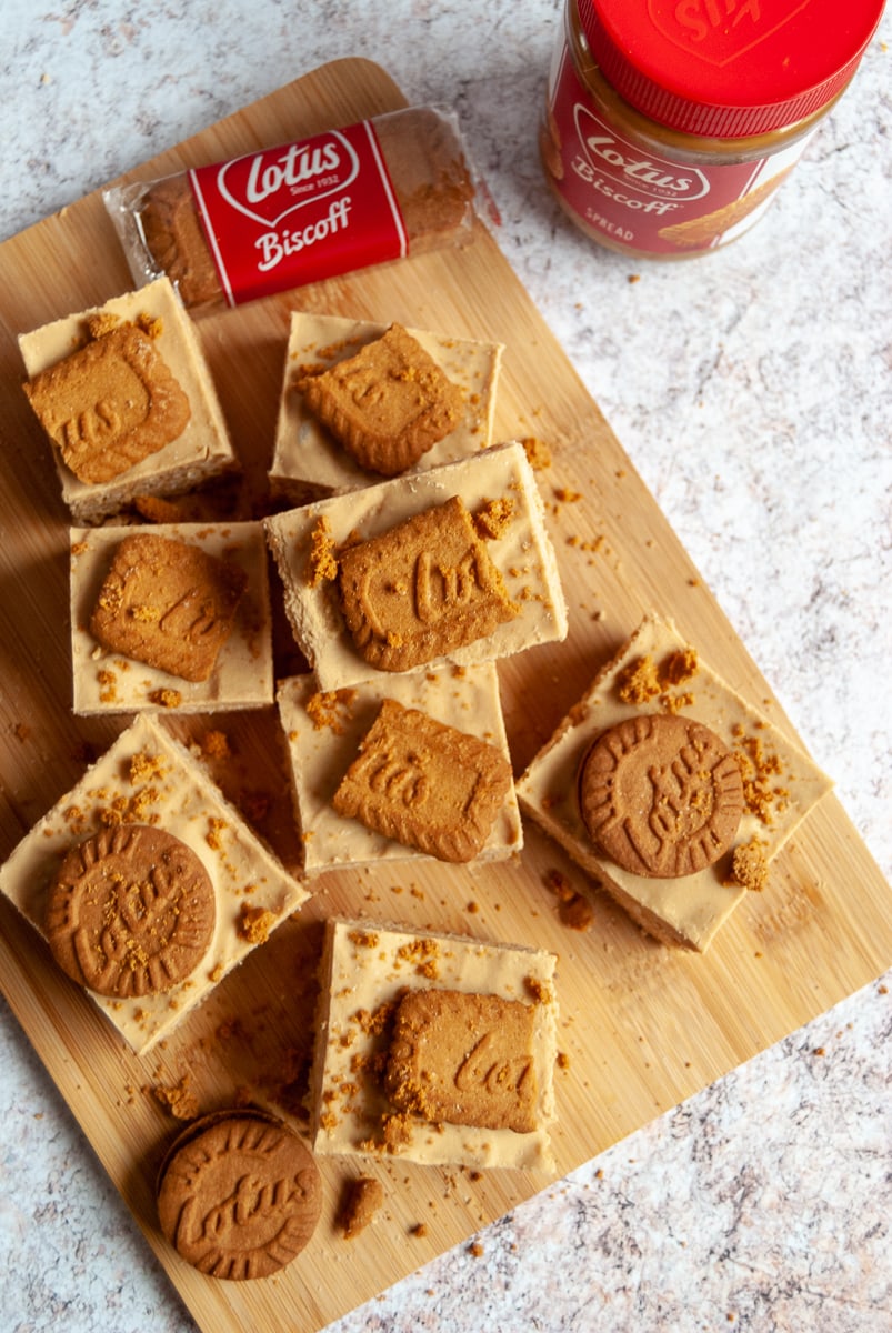 Biscoff marshmallow squares topped with biscoff cookies on a wooden board. A jar of biscoff spread can be partially seen