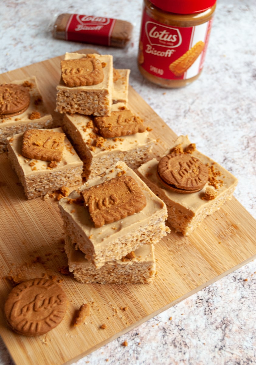 squares of biscoff rice krispie treats topped with biscoff cookies on a wooden board with a jar of Biscoff spread in the background