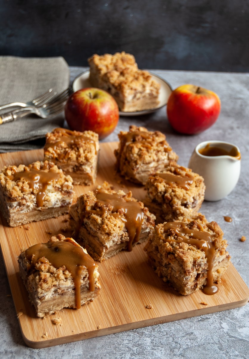 Apple cheesecake bars covered in caramel sauce on a wooden board, a small white jug of caramel sauce, a beige linen napkin and two red apples