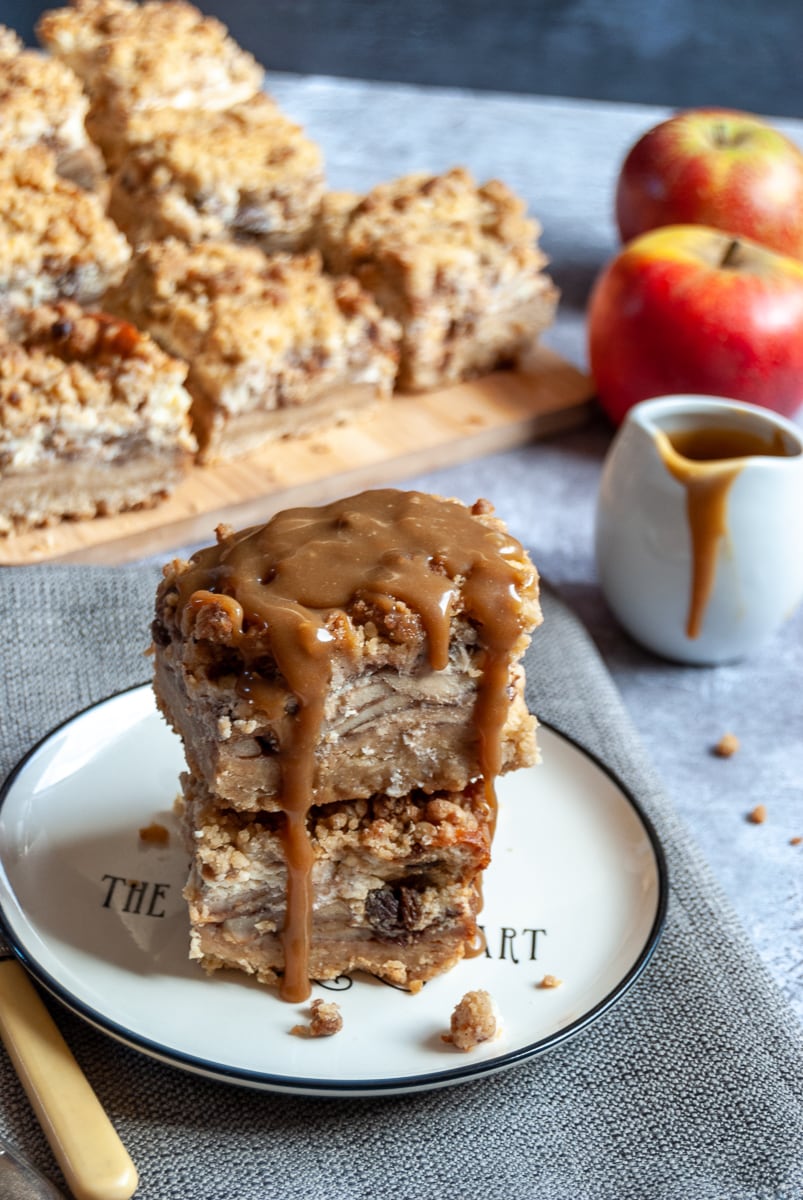 two apple cheesecake bars drizzled with caramel sauce on a white and black plate with a grey napkin and a yellow fork, a small white jug of caramel sauce, two red apples and more cheesecake bars on a wooden board in the background.