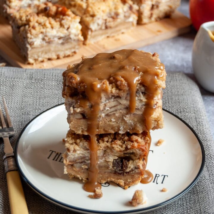 Two apple cheesecake bars covered in caramel sauce on a white and black plate, a yellow fork and more apple cheesecake bars on a wooden board in the background