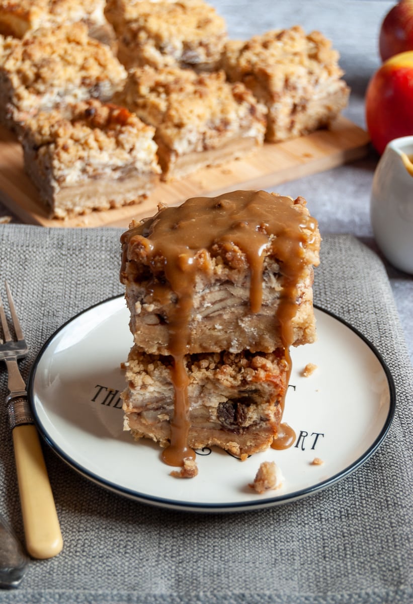 Two apple cheesecake bars covered in caramel sauce on a white and black plate, a yellow fork and more apple cheesecake bars on a wooden board in the background