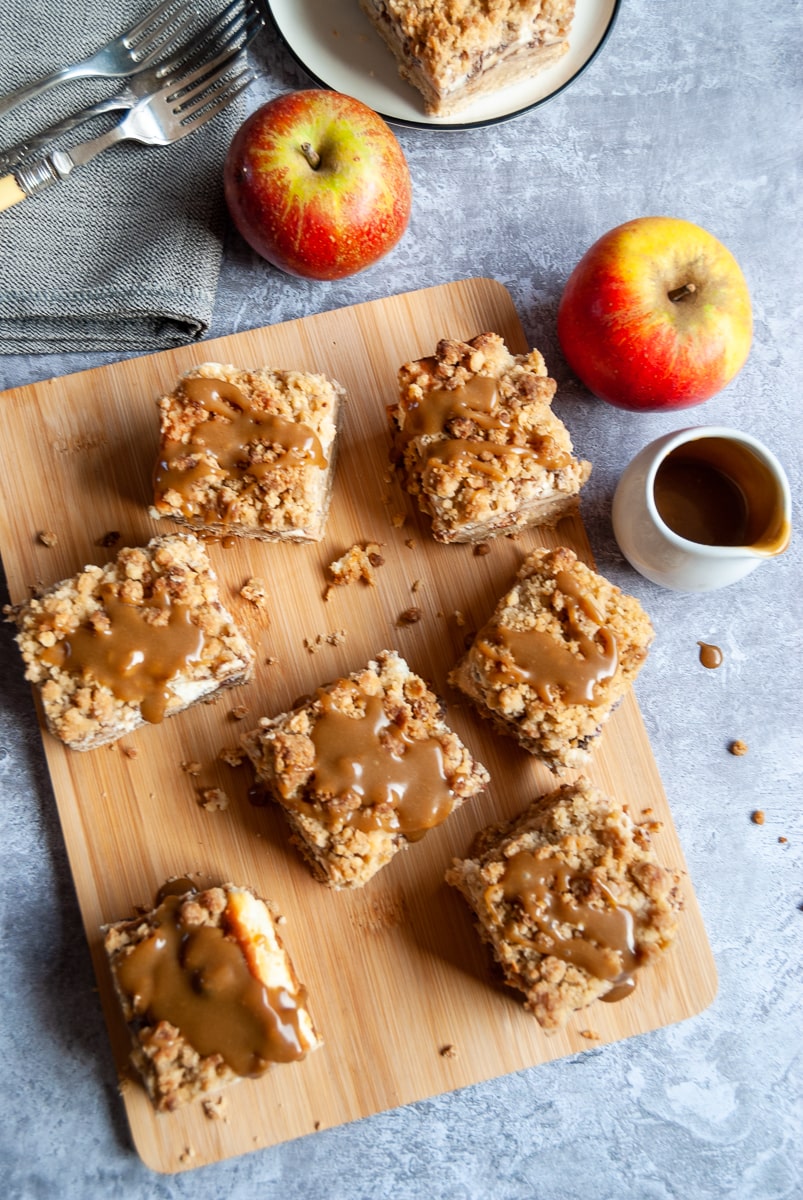 A flat lay photo of apple cheesecake bars with caramel sauce on a wooden board, a small white jug of caramel sauce and two red apples