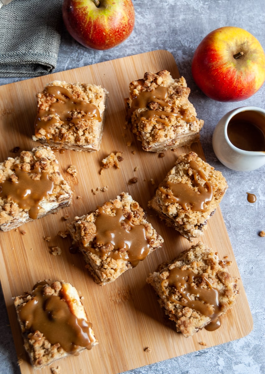 apple cheesecake bars with a crumble topping and caramel sauce on a wooden board with a small white jug of caramel sauce and two red apples