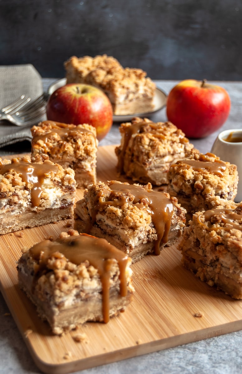 apple cheesecake bars covered in caramel sauce on a wooden board, a linen napkin and two red apples in the background