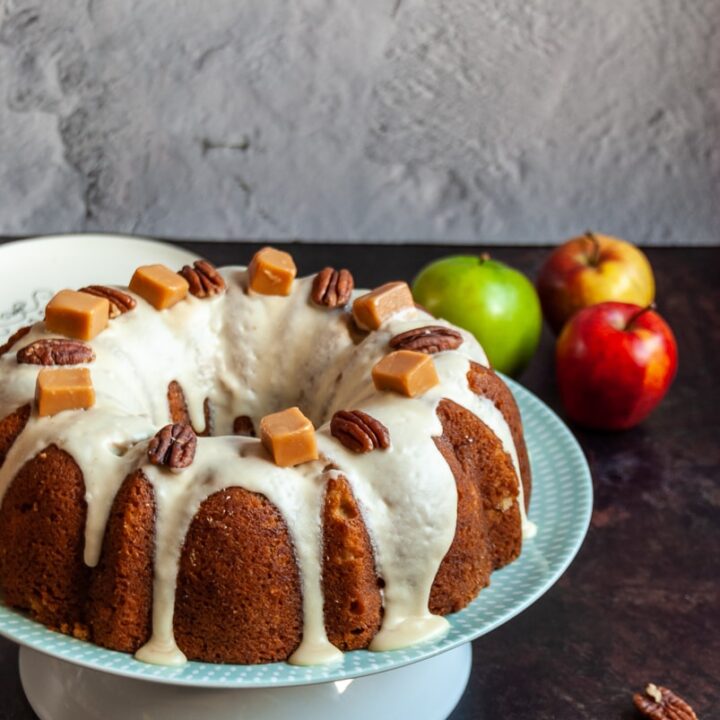 an apple bundt cake covered in caramel icing and topped with pecan nuts and fudge pieces on a white and green cake stand, apples in the background and nuts and fudge pieces scattered around the cake