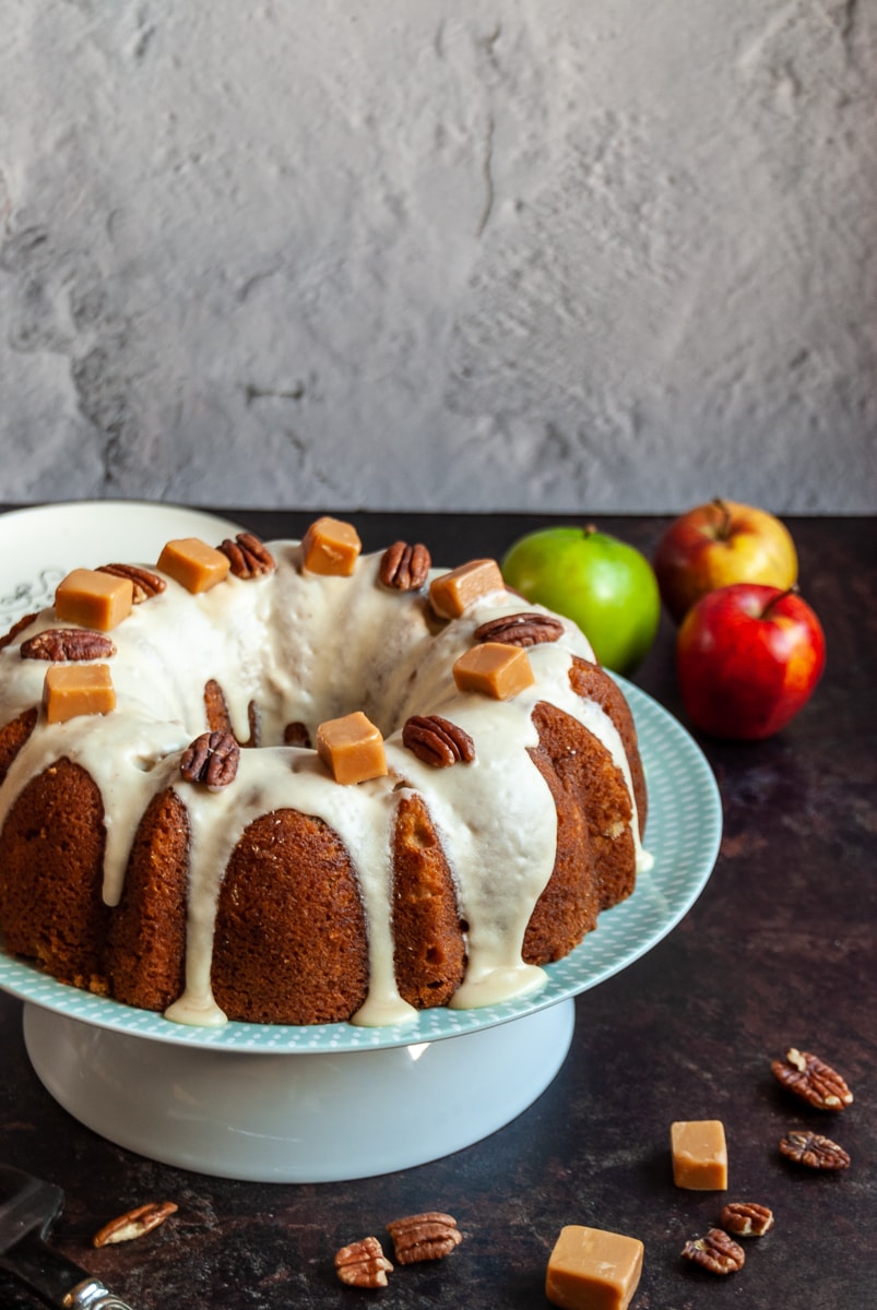 an apple bundt cake covered in caramel icing and topped with pecan nuts and fudge pieces on a white and green cake stand, apples in the background and nuts and fudge pieces scattered around the cake