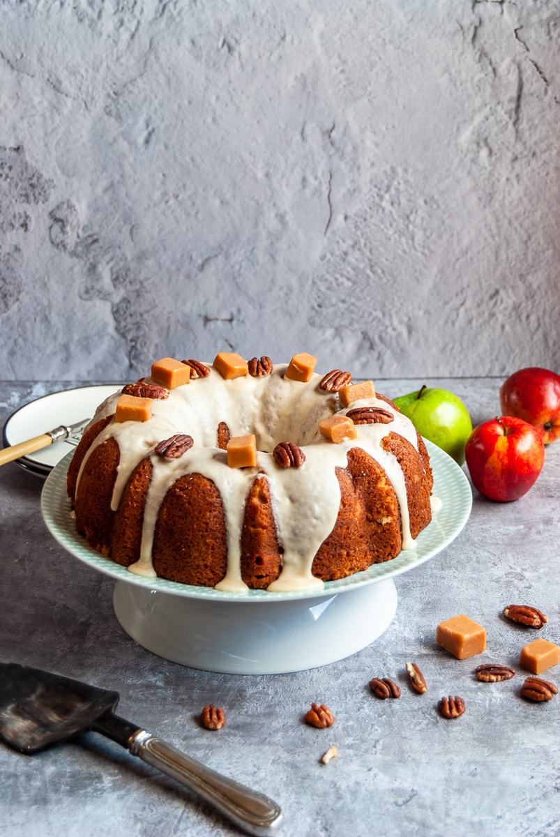 an apple bundt cake covered in caramel glaze and topped with pecan nuts and fudge pieces on a white and green cake stand, small plates and a fork in the background with apples, pecan nuts and fudge pieces scattered around the cake