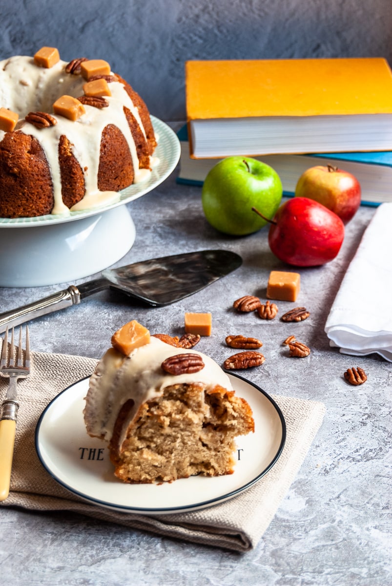 a slice of apple bundt cake covered in caramel glaze and topped with pecan nuts and a fudge chunk on a white and black plate and a folded beige napkin with a fork, a large apple bundt cake on a cake stand in the background with a vintage cake slice, apples, pecan nuts and fudge chunks scattered around and two books