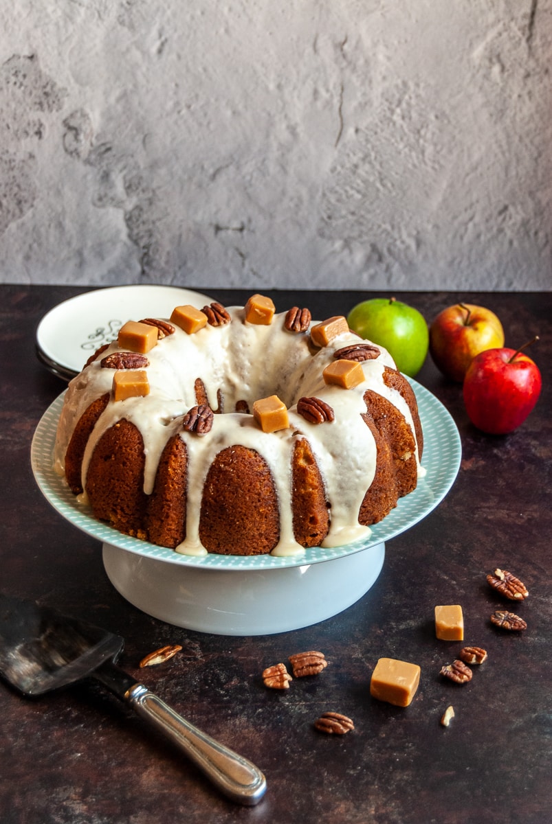 an apple bundt cake covered in caramel icing and topped with pecan nuts and fudge pieces on a white and green plate. small serving plates and apples in the background and pecans and fudge pieces are scattered around the picture