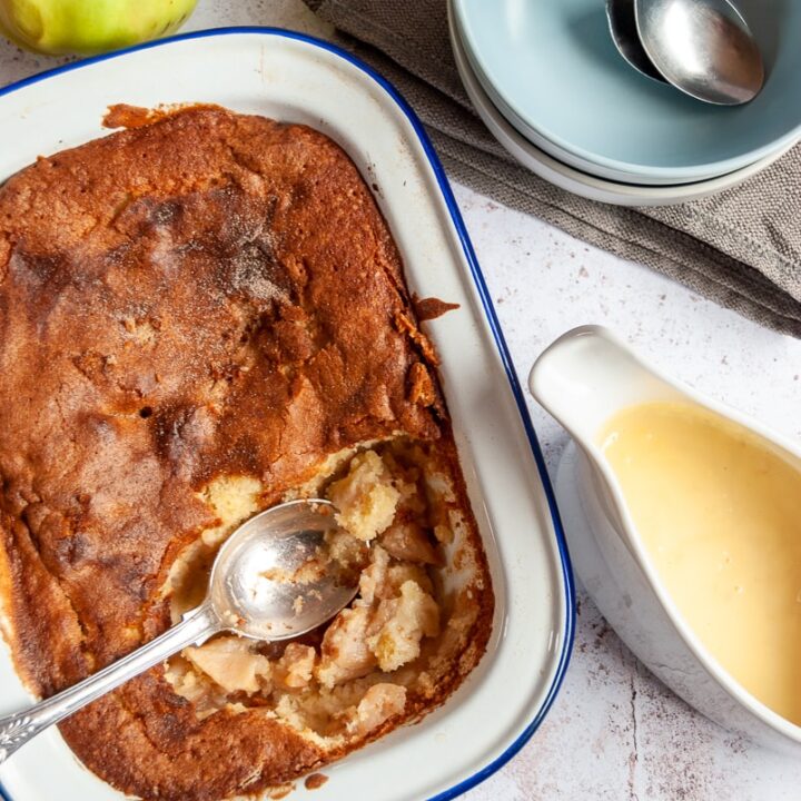 an apple sponge pudding with a spoon in a blue and white baking tin, a white jug of custard, two bowls with spoons and green apples.