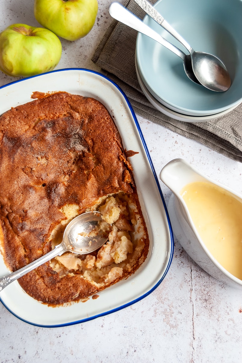 an apple sponge pudding with a spoon in a blue and white baking tin, a white jug of custard, two bowls with spoons and green apples.