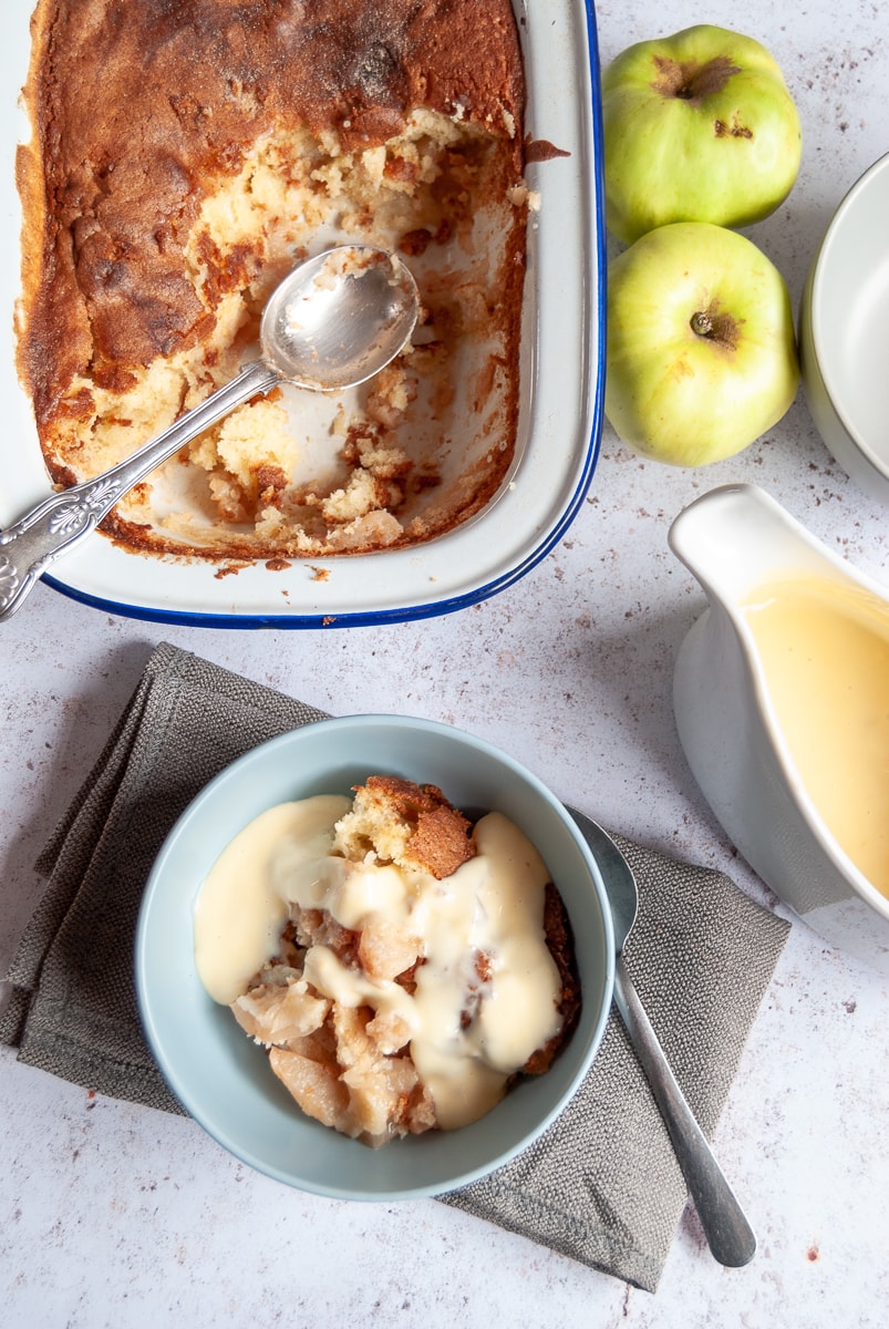 A flat lay image of a blue bowl of apple sponge with custard on a grey napkin and a spoon, a white jug of custard, a dish of apple sponge pudding and two green apples