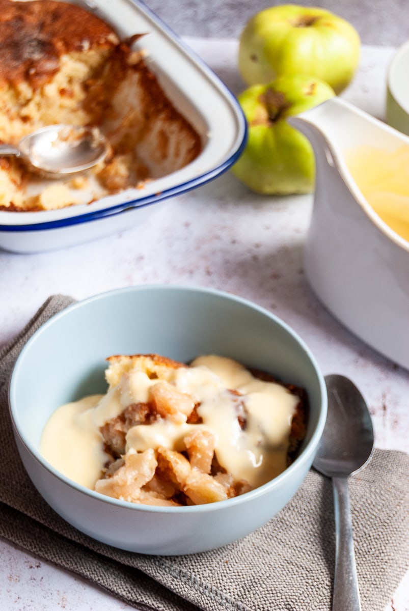 a blue of apple sponge pudding with custard on a grey napkin with a spoon sitting beside the bowl, a white jug of custard and more apple pudding in a large blue and white baking tin 