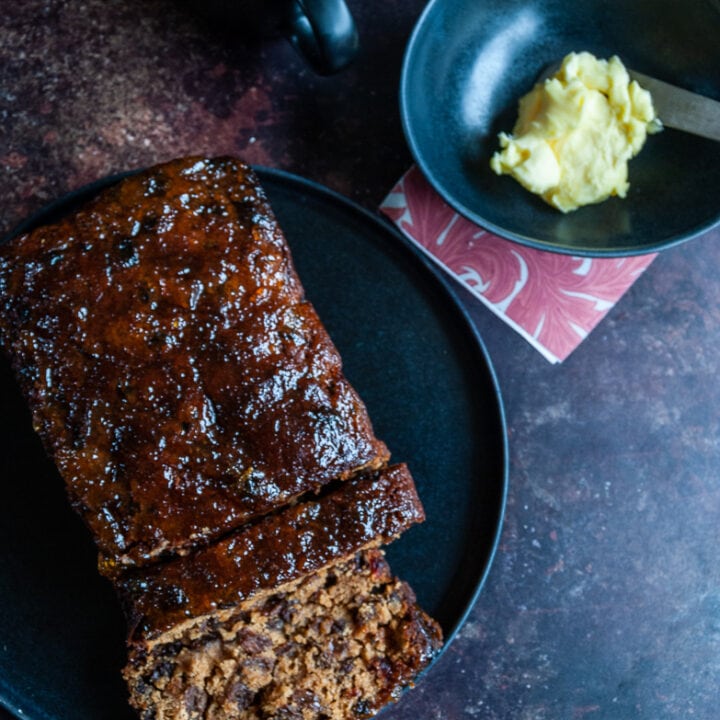 a fruit loaf on a black plate, a cup of tea in a black mug and a pot of butter with a white handled knife