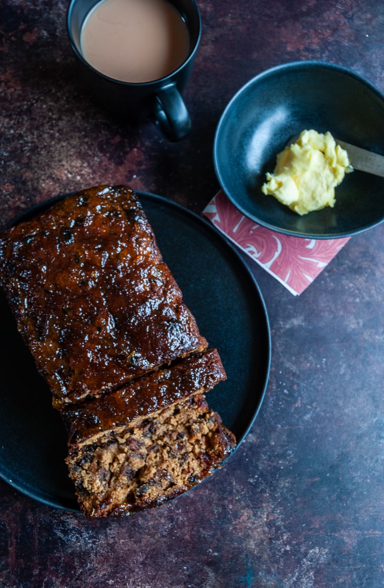 a fruit loaf on a black plate, a cup of tea in a black mug and a pot of butter with a white handled knife