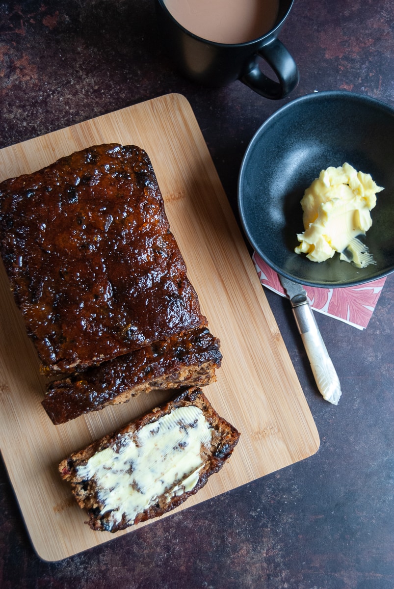 a fruit loaf cake on a wooden board and a buttered slice of the cake, a pot of butter and a black mug of tea