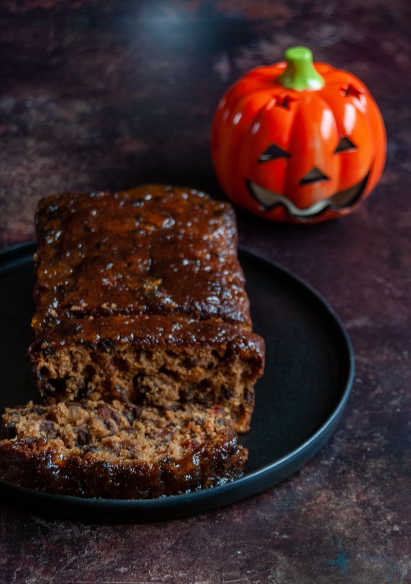 A glazed fruit loaf on a black serving plate and a small ornamental pumpkin in the background
