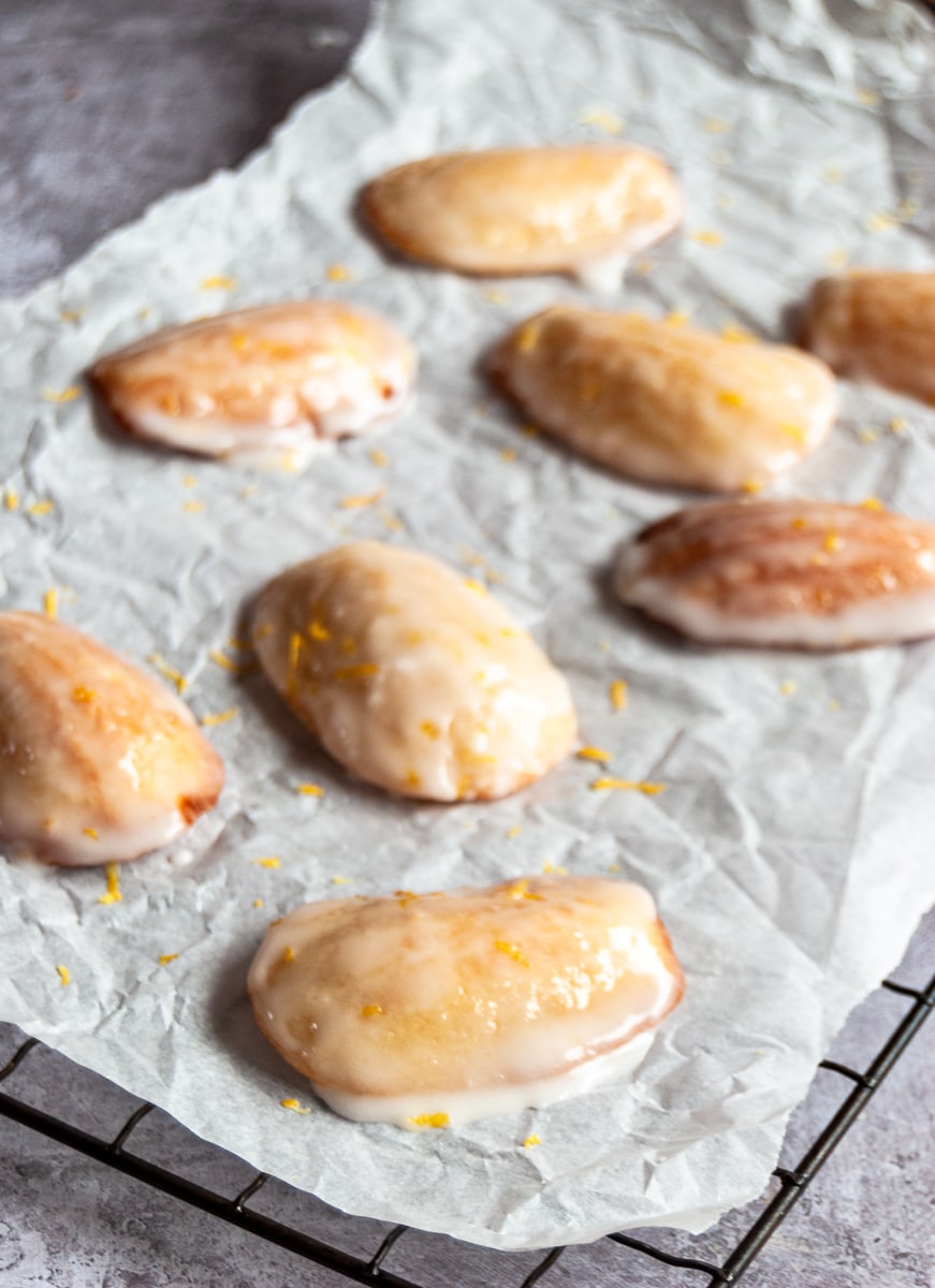 a close up picture of madeleines with a lemon icing glaze and scattered with lemon zest on a piece of baking parchment