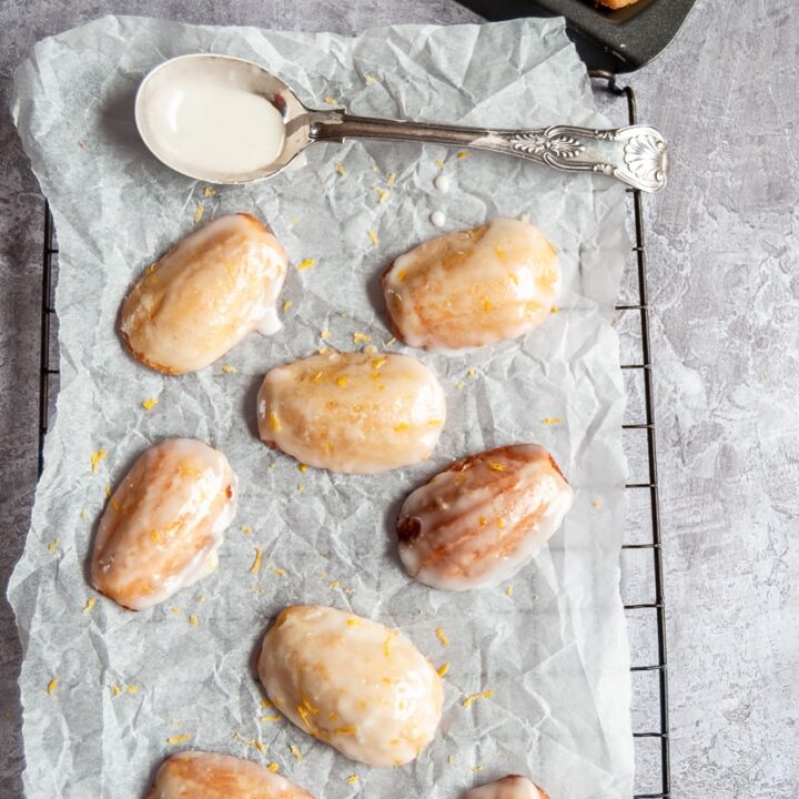 eight madeleines with a lemon icing and grated lemon zest on a piece of baking parchment/wire rack with a spoon and a madeleine pan can be partially seen.