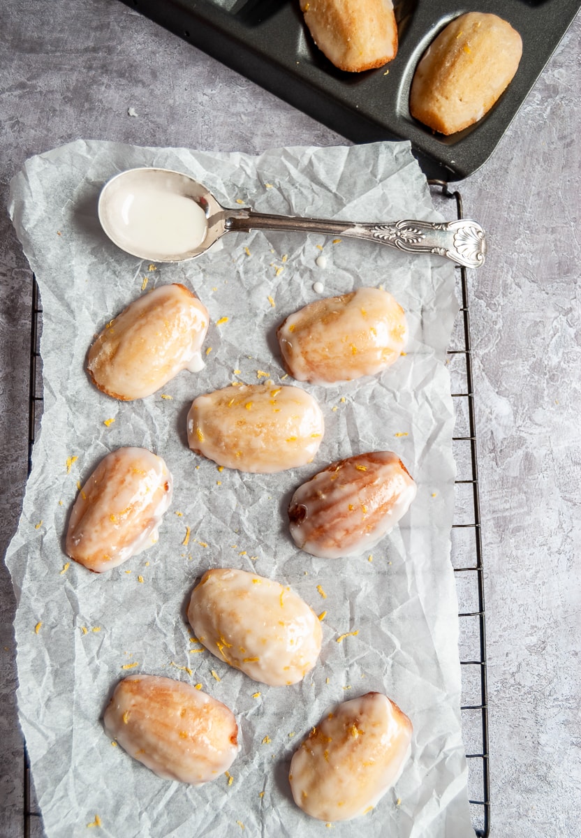 eight madeleines with a lemon icing and grated lemon zest on a piece of baking parchment/wire rack with a spoon and a madeleine pan can be partially seen.