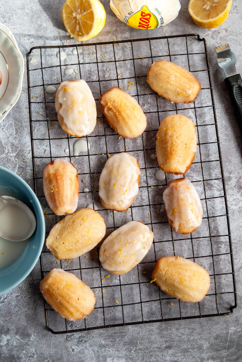 Madeleines topped with lemon icing glaze and sprinkled with lemon zest on a black wire cooling rack, fresh lemons, a blue bowl with lemon icing and a spoon and a black lemon zester.