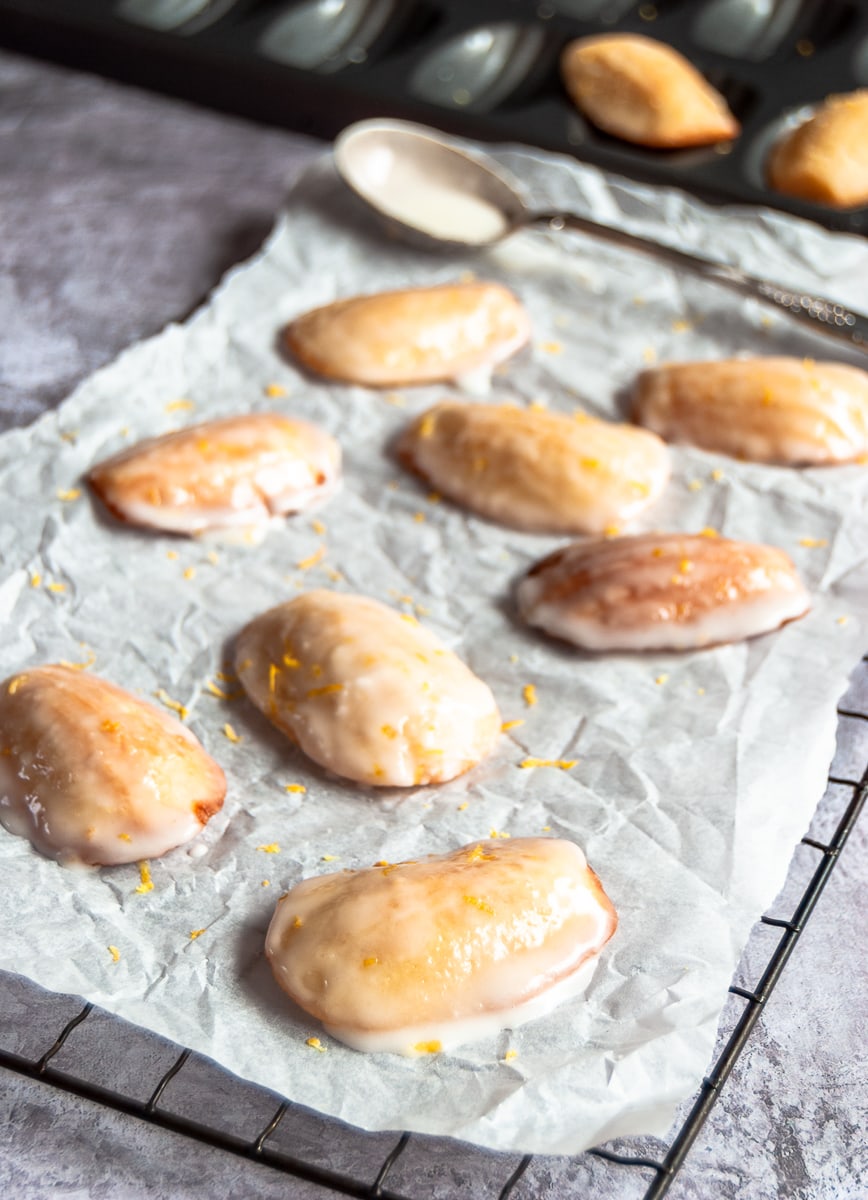 a close up photograph of lemon madeleines topped with a lemon icing glaze and sprinkled with lemon zest on a piece of baking parchment and a black cooling rack with a spoon and a madeleine pan in the background.
