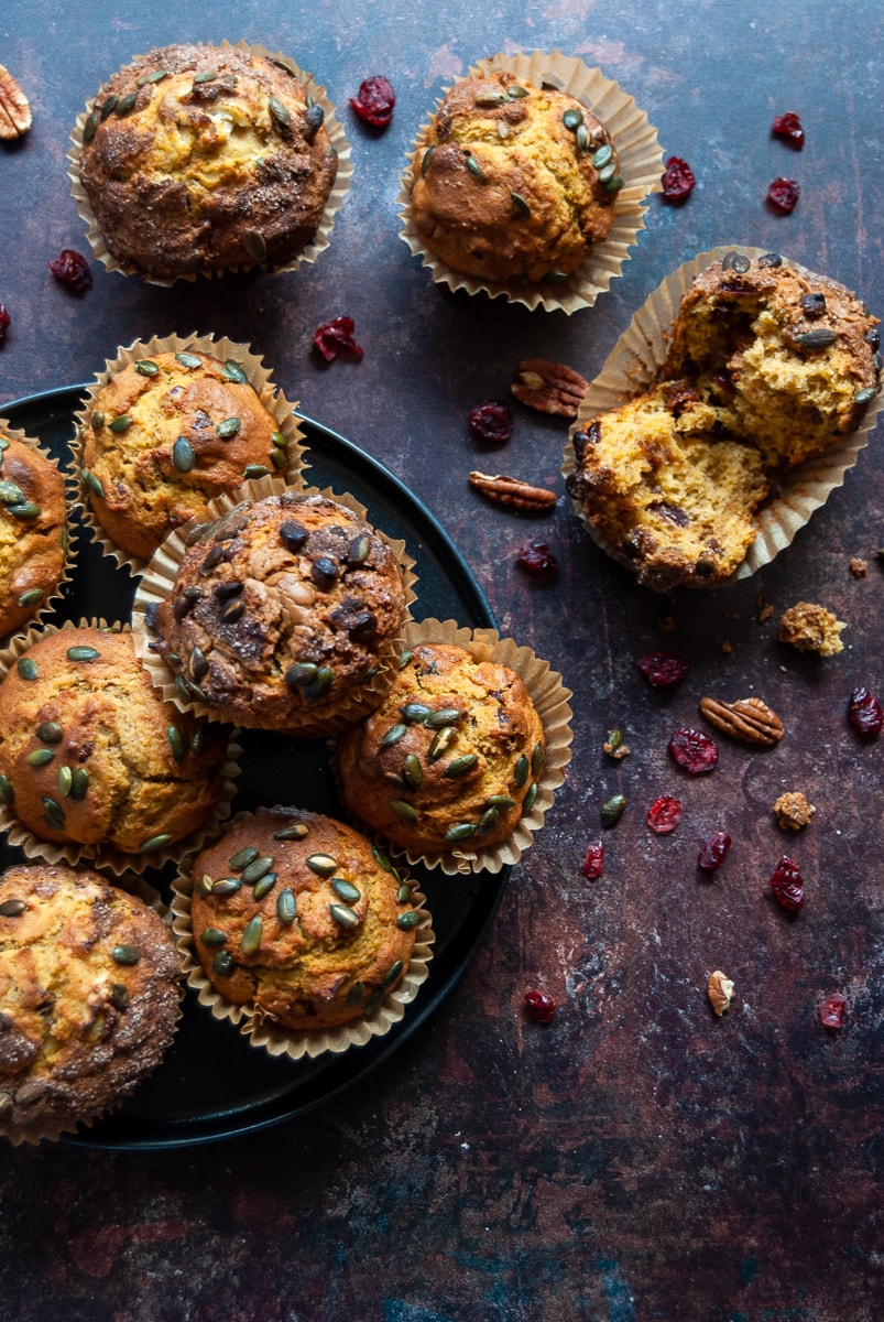 A black plate of pumpkin muffins with cranberries, pumpkin seeds and cinnamon sugar on a backdrop of Autumnal colours with cranberries and pumpkin seed scattered around the muffins
