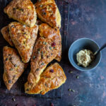 A flat lay photo of pumpkin scones sprinkled with pumpkin seeds and cranberries on a black wire rack and a grey pot of maple butter