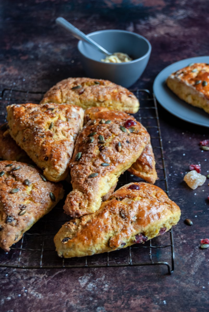 a batch of pumpkin scones with dried cranberries and pumpkin seeds on a black wire rack. A scone on a grey plate and a small grey pot of maple butter can be seen in the background.