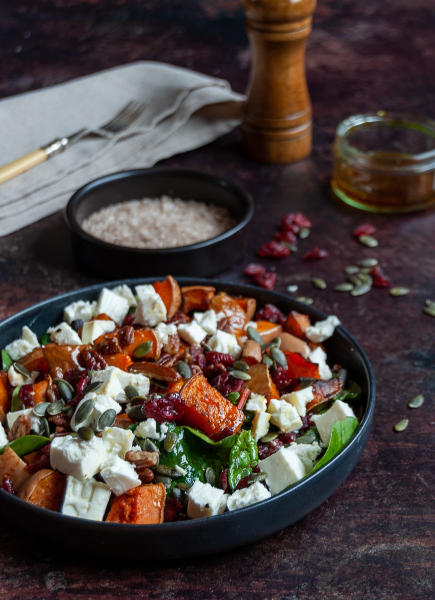 a black stoneware bowl of butternut squash, cranberry, feta cheese and spinach salad, a small black pot of sea salt, a beige napkin with a fork, a wooden pepper mill and a small pot of salad dressing.