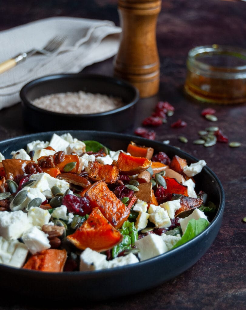 a black stoneware dish with a butternut squash, cranberry, pecan and feta cheese salad, a small black pot of sea salt, a wooden pepper mill, a small jar of salad dressing and a beige napkin with a fork.