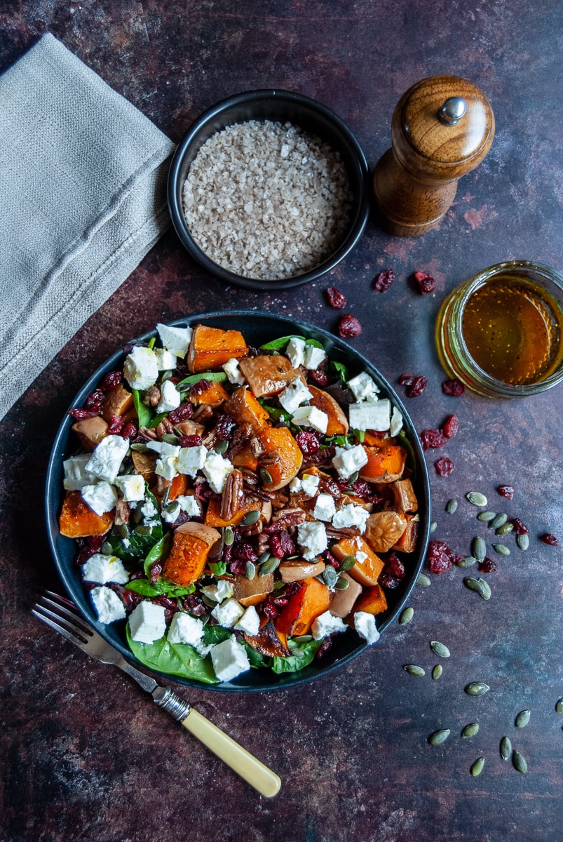 a flat lay photo of a bowl of butternut squash, pecan, feta cheese and cranberry salad with spinach leaves, a small black pot of sea salt, a wooden pepper mill, a beige napkin, a small jar of salad dressing and a yellow handled fork.