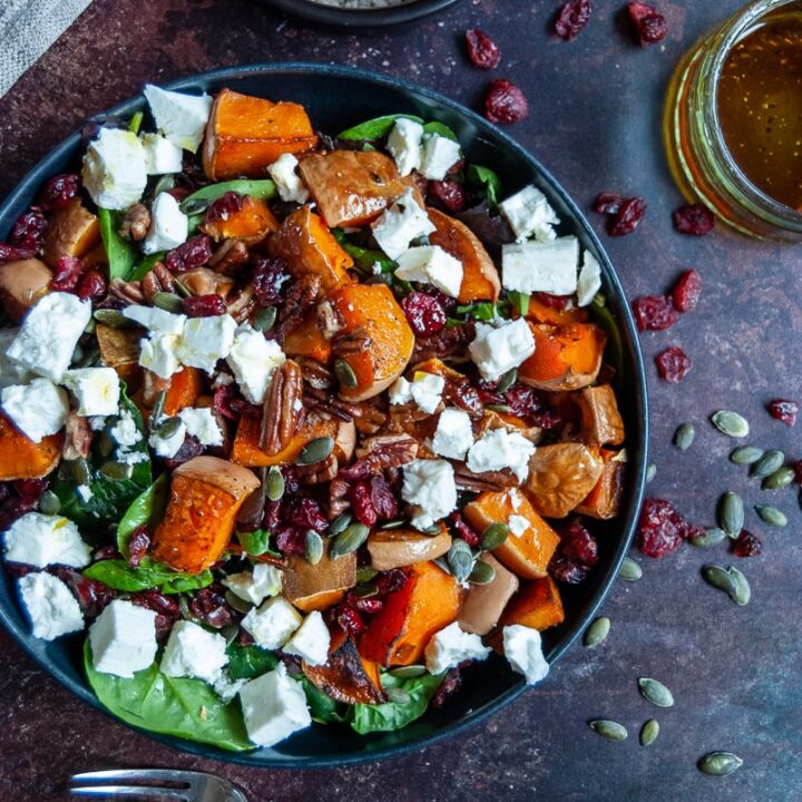 a flat lay photo of a bowl of butternut squash, pecan, feta cheese and cranberry salad with spinach leaves, a small black pot of sea salt, a small jar of salad dressing and a yellow handled fork. A wooden pepper mill and beige napkin can be partially seen.