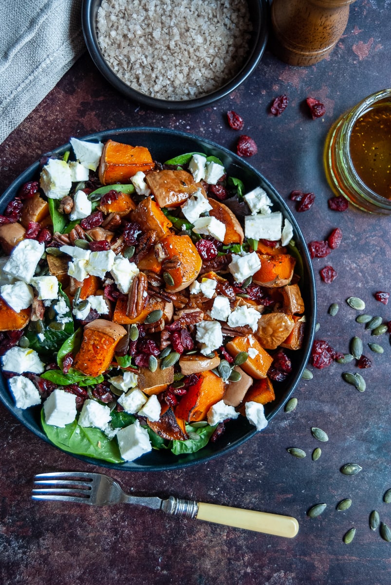 a flat lay photo of a bowl of butternut squash, pecan, feta cheese and cranberry salad with spinach leaves, a small black pot of sea salt, a small jar of salad dressing and a yellow handled fork. A wooden pepper mill and beige napkin can be partially seen.