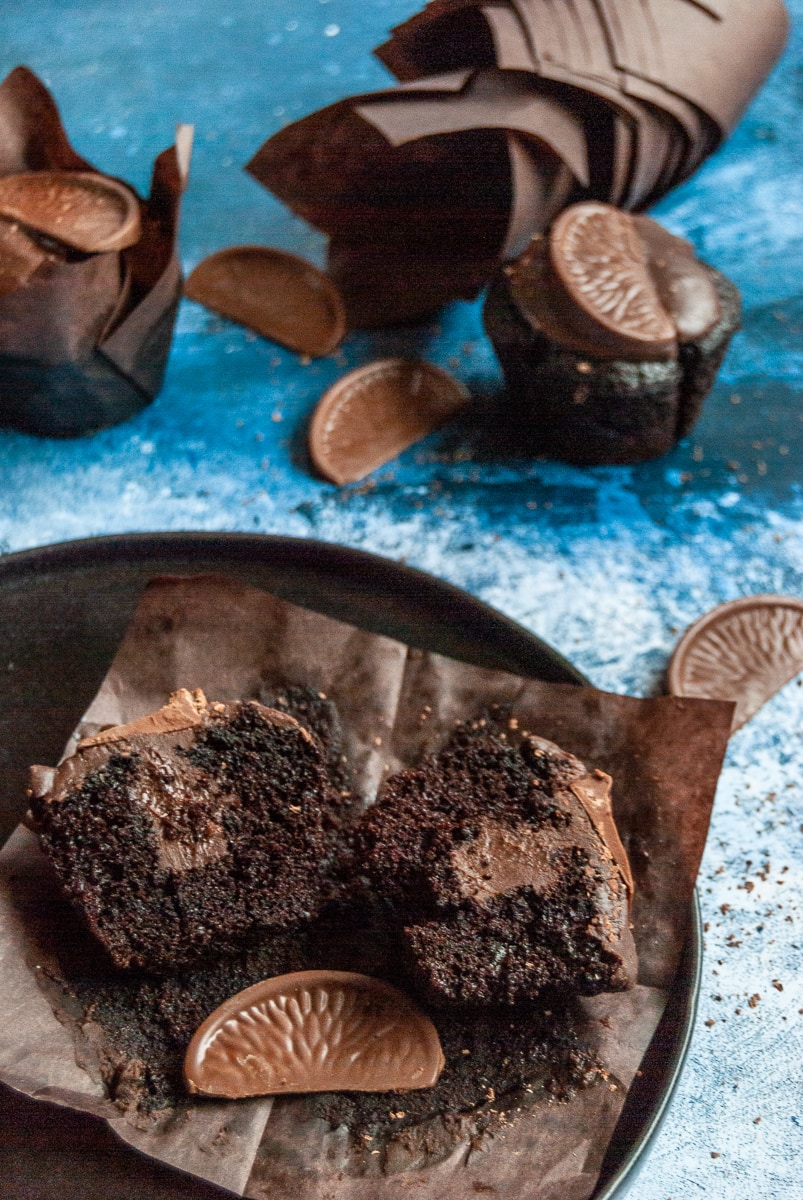 a chocolate orange muffin on a black plate cut in half to reveal a chocolate orange ganache filling. More chocolate muffins topped with chocolate ganache and a terry's chocolate orange segment sit above the plate.