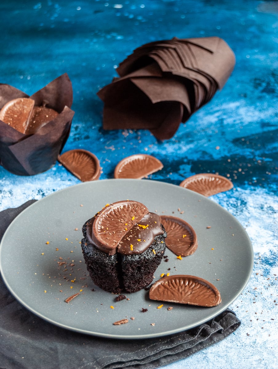 A chocolate cupcake topped with chocolate ganache and a terry's chocolate orange segment on a grey plate and black napkin. More cupcakes and chocolate oranges can be seen on a blue backdrop.