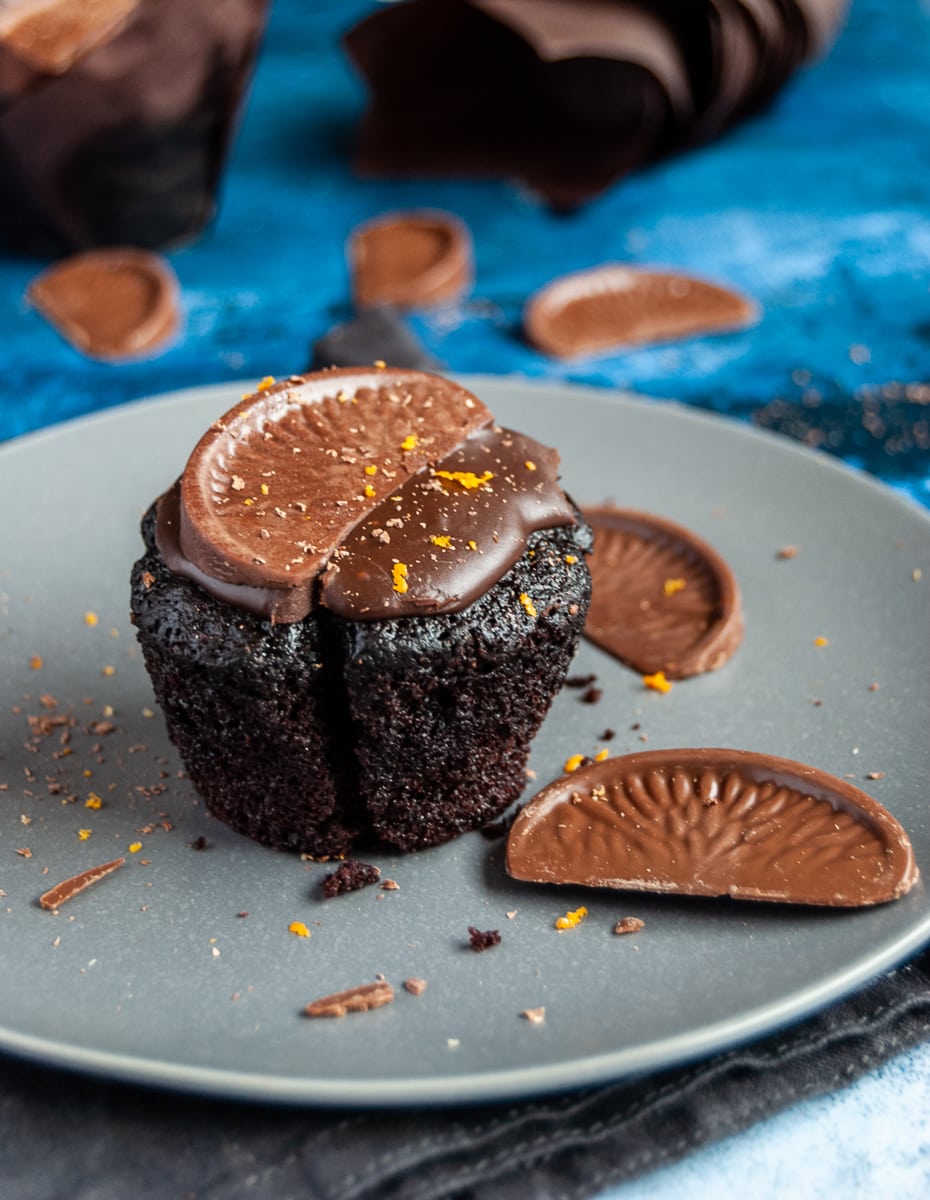 a chocolate orange muffin topped with chocolate ganache and a terry's chocolate orange segment on a grey plate with more chocolate orange segments. 